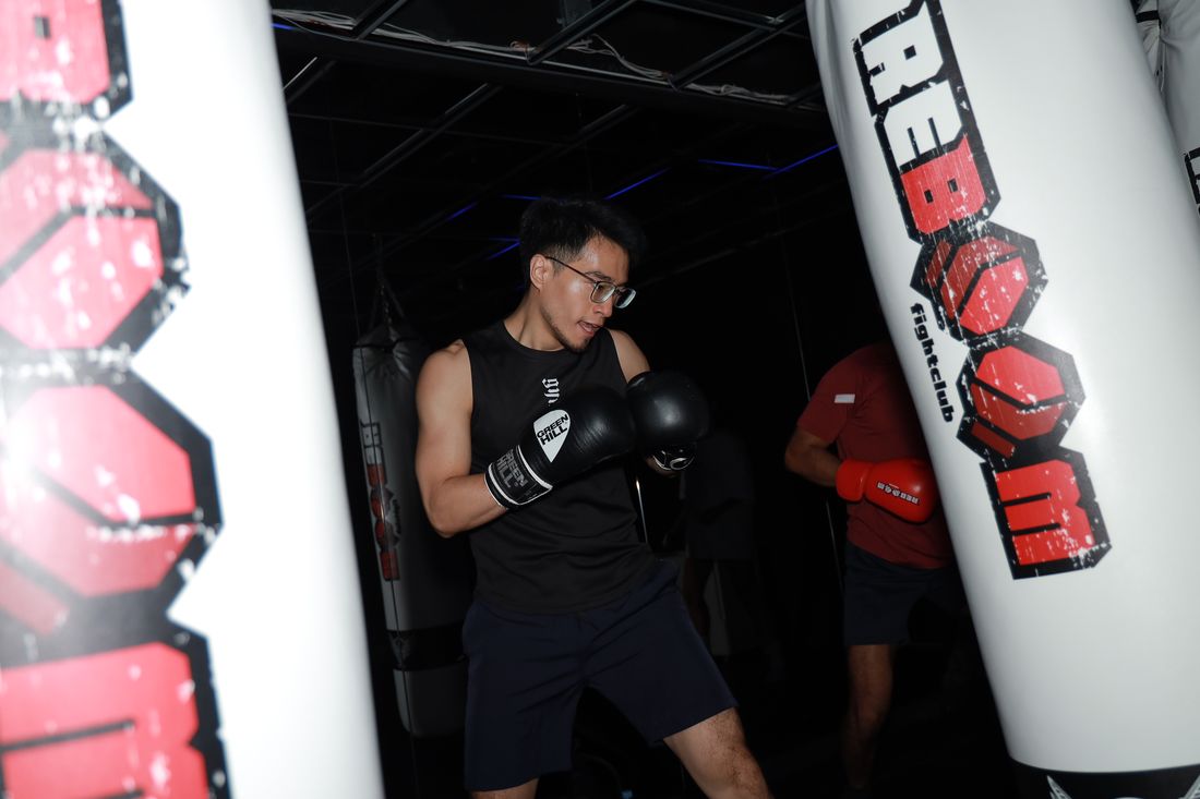 一位戴眼鏡的男生穿黑色背心,戴著拳擊手套在兩顆沙包之間出拳