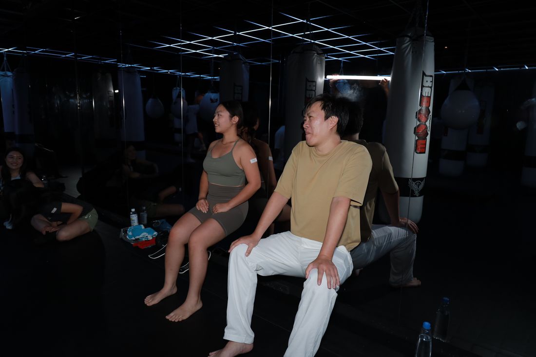 兩位參加者背靠著牆做靠牆半蹲,表情吃力,旁邊有人坐在地上看著